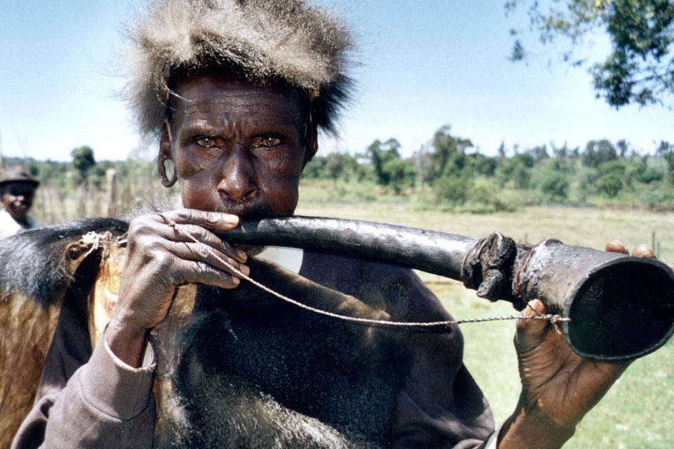 A photograph of an indigenous man blowing into a crude horn