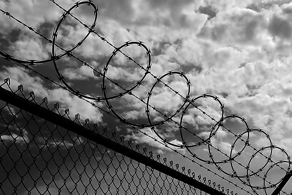 A black and white photograph looking up at the sky through a chain link fence topped with barbed wire