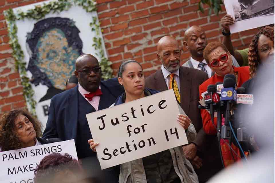 A photograph of a number of African Americans at a press conference. The woman in the center holds a sign that reads, &quot;Justice for Section 14&quot;