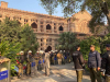 A photograph of the exterior of a university building. Police line the walk leading in, fully suited in riot gear.