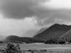 A moody black and white photo of a landscape at the foot of a tree covered mountain