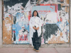 A photograph of a dark skinned woman standing in front of a urban wall