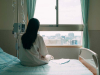 A photograph of a woman in a hospital gown seated away from the camera, looking out a nearby window