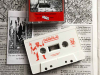 A white cassette with the world Kobold printed on it in red. The cassette lays on a garishly illustrated text