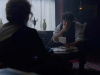 A figure with their back to the viewer looks on as a seated man bemusedly looks at a piece of paper