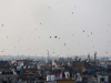 A distant shot of a cityscape. Kites dot the sky are rising into the air.