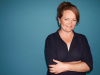 A photograph of Dorthe Nors, who smiles at the viewer, arms folded, against a dark blue background