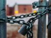 A close-up on a padlock on a chain holding a link fence closed. A building can be made out in the unfocused background