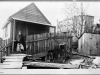 An historical black and white photograph of an indigenous woman standing on the porch of a simple home newly constructed from lumber