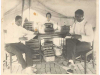 Two African American men seated at a table in a tent. Law books are stacked on the table. A woman stands at the entrance to the tent in the background.