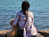 A photograph of a dreadlocked figure sitting with a dog looking out over a body of water
