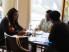 Three people sit around a table writing with a window illuminating the table