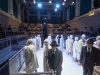 A photograph showing male and female actors dressed in either white trenchcoats or black suits. A socially distanced audience can be seen on bleachers surrounding the stage.