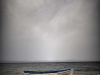 Orange lifejackets piled up on the shore of an island just next to a boat moored nearby. The open ocean and a dark sky looms in the background.