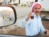 A man wearing a red and white headwrap smiles as he offers the viewer, who is inside of a car, a small, clear glass of tea