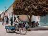 A photograph of a man riding a bicycle, pulling a small blue wagon, down a quiet street with people sitting and standing on the sidewalk. A tree offers shade to a few people sitting beneath it