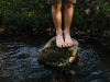 A pair of childish legs stand perched on a mossy rock in a brook