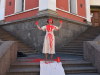 A photograph of a woman dressed in white standing at the intersection of two sets of stairs. She is splattered with red paint.