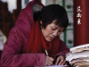 A photograph of a woman in a heavy red parka, hunched over a desk, writing