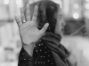 A black and white photograph of a woman, whose head is covered by a scarf, pressing her hand to a damp window between herself and the viewer. Her gaze is away from the viewer.