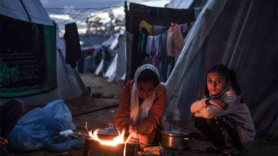 Three figures sit around a small fire outside of a canvas tent