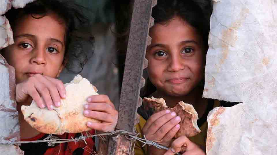 Two children eating bread peer through a crack in a ruined structure
