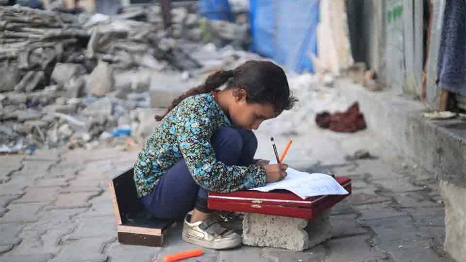 A young girl kneels in the rubble to use a displaced stone as a desk to work on schoolwork