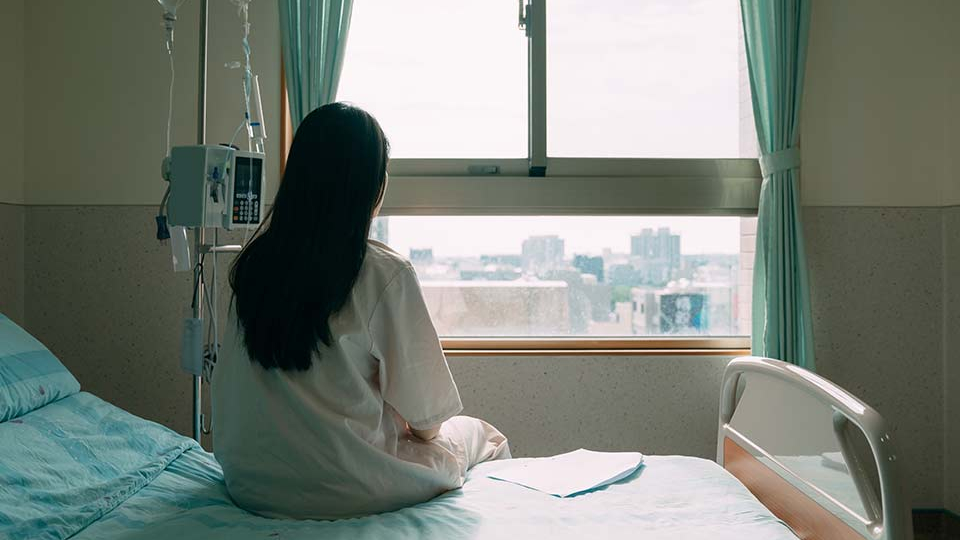 A photograph of a woman in a hospital gown seated away from the camera, looking out a nearby window