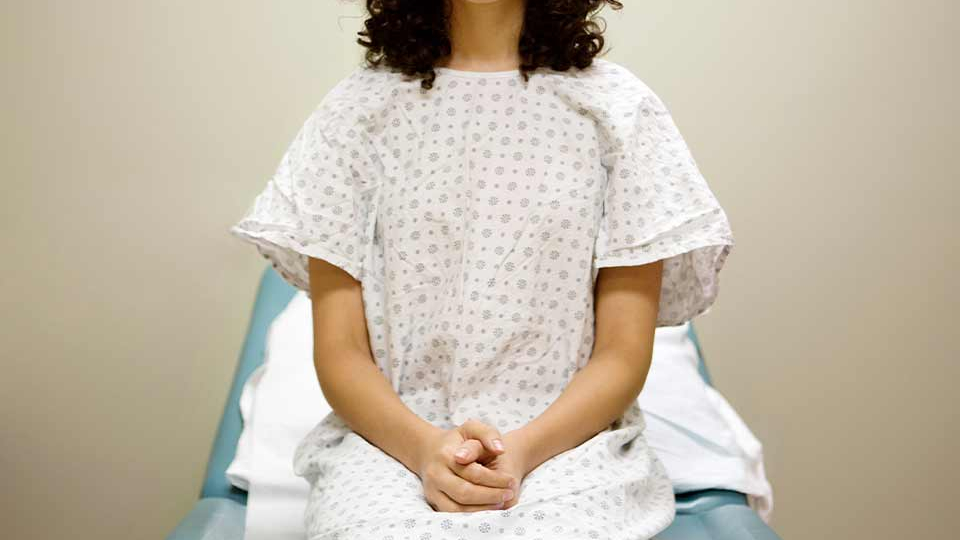 A woman in a hospital gown sitting on an examination table with her hands folded in her lap