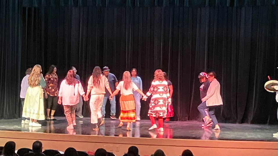 A group of people dance on a bare stage while holding hands
