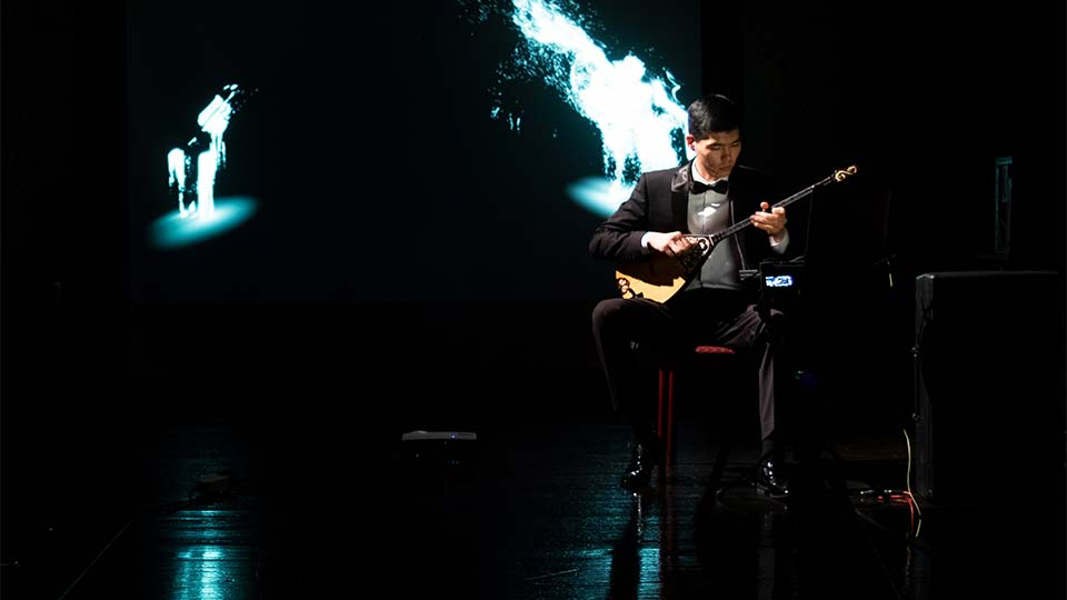 A man seated on a mostly darkened stage playing a saz