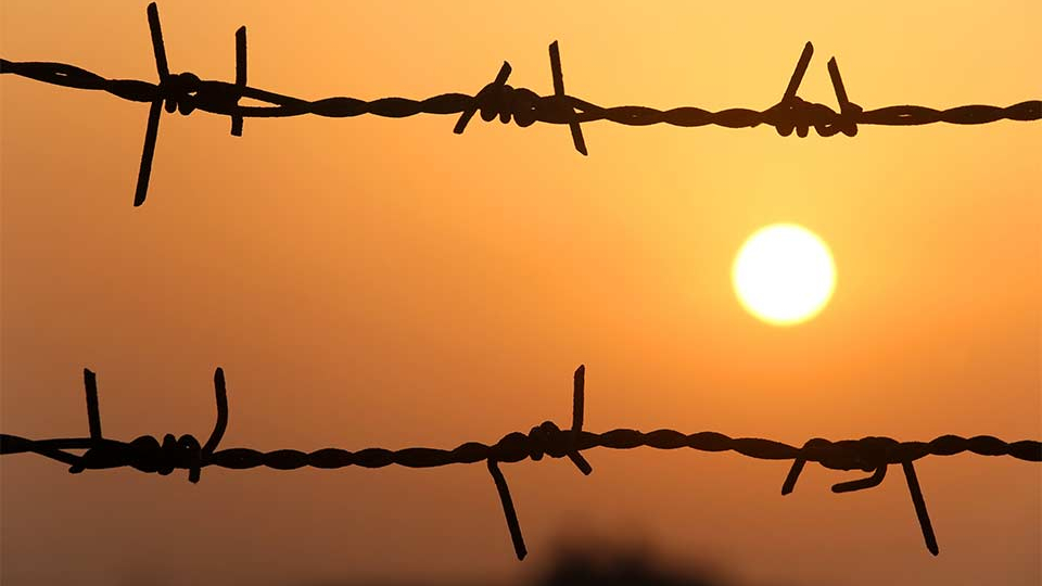 A bleary orange sun is setting, as seen through a barbed wire fence.
