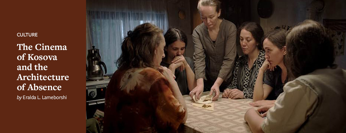 A group of women seated around a table. One woman, who is standing, appears to unfolding a cloth to reveal gold within