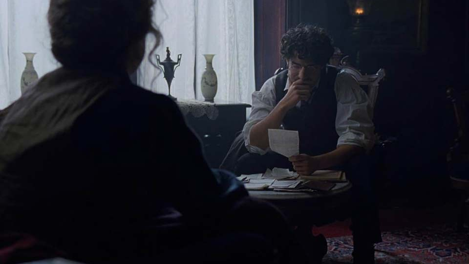 A figure with their back to the viewer looks on as a seated man bemusedly looks at a piece of paper