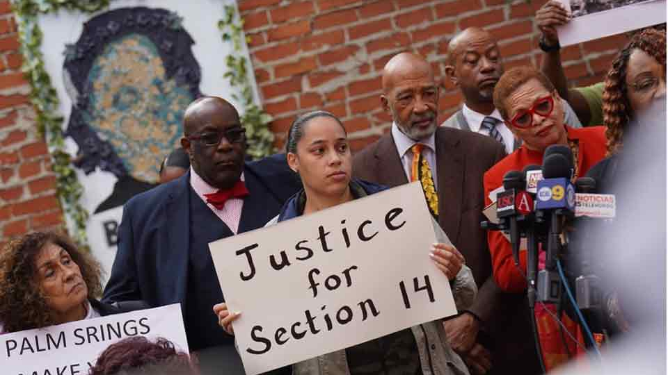 A photograph of a number of African Americans at a press conference. The woman in the center holds a sign that reads, "Justice for Section 14"
