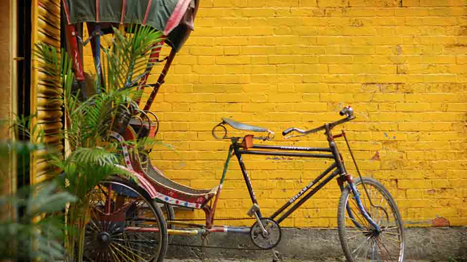 A bicycle connected to a carriage, parked in front of a mustard-colored brick wall