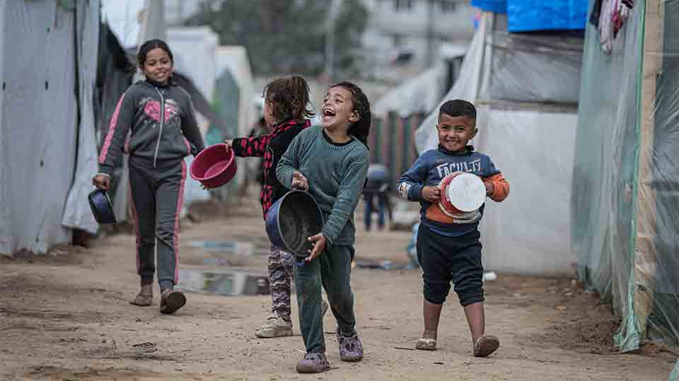 A group of refugee children carrying water basins, smiling as they walk together through a tent city