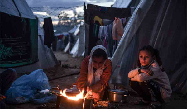 Three figures sit around a small fire outside of a canvas tent