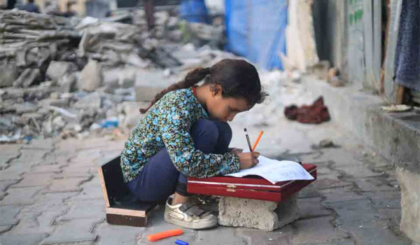 A young girl kneels in the rubble to use a displaced stone as a desk to work on schoolwork
