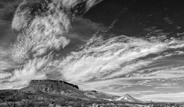 A black and white photograph of a cloud streaked sky above a mesa and the scrublands below