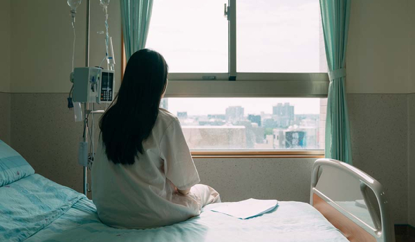 A photograph of a woman in a hospital gown seated away from the camera, looking out a nearby window