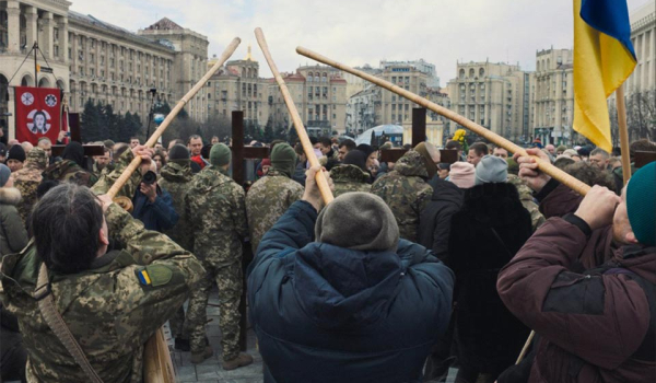 A photograph of a group of three people holding long wooden poles to their face in a crowd of protesters