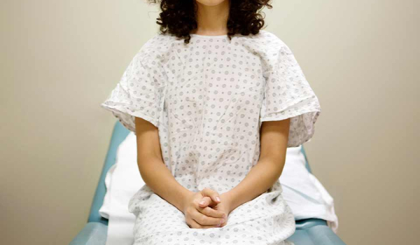 A woman in a hospital gown sitting on an examination table with her hands folded in her lap