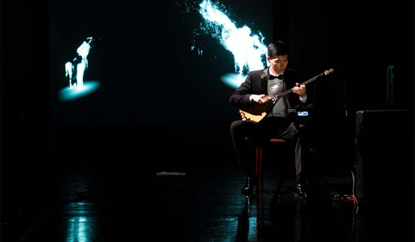 A man seated on a mostly darkened stage playing a saz