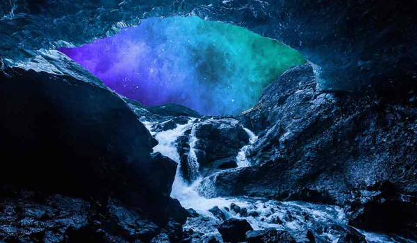 A river flows into the mouth of a darkened cave over rocks, creating a froth. The sky outside the cave is lit up with an aurora borealis