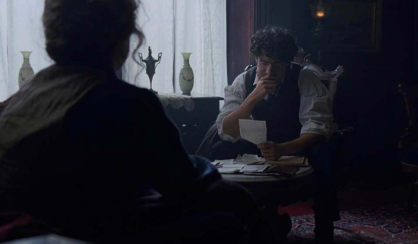 A figure with their back to the viewer looks on as a seated man bemusedly looks at a piece of paper