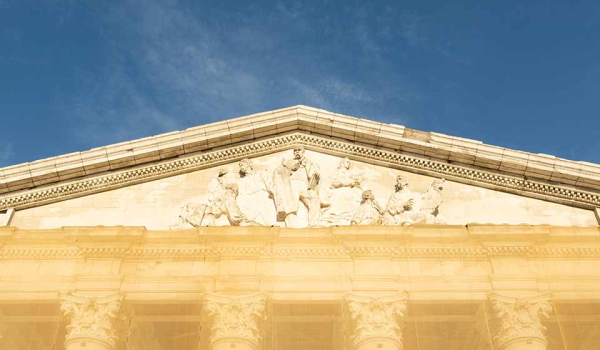 A photograph of a detail near the roofline of an aged marble building. The area has a relief sculpted into including several historical and mythological figures, including Joseph Conrad