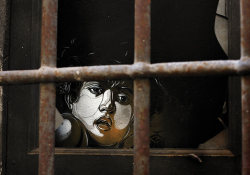 The drawn image of a human face peers from within a darkened room through iron bars