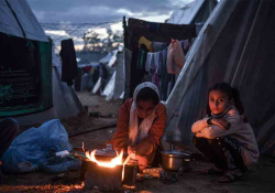 Three figures sit around a small fire outside of a canvas tent