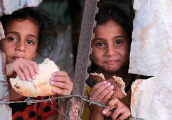 Two children eating bread peer through a crack in a ruined structure